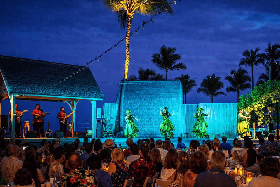Hula dancers and musicians on the outdoor Island Breeze Luau stage, with guests seated in front at dusk